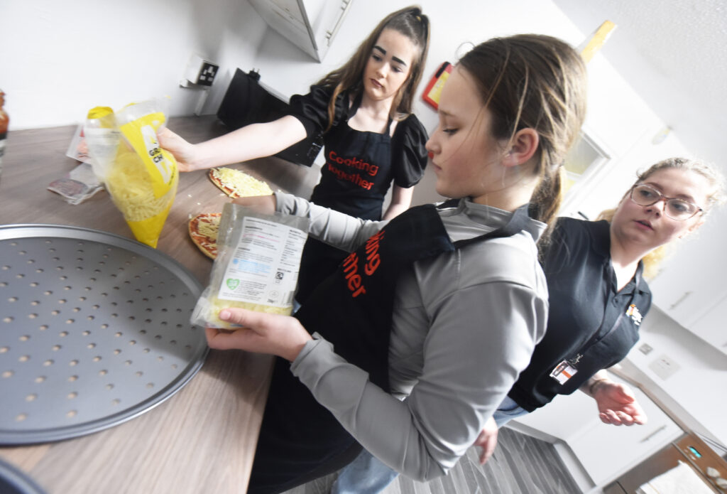 Two younger individuals wear black aprons with "Cooking Together". One is holding a bag of grated cheese over a pizza base on the counter, while the other looks on.