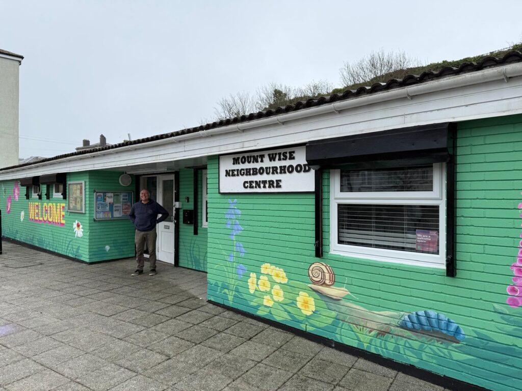 Simon standing outside the freshly painted Mount Wise Neighbourhood Centre in Plymouth, its exterior walls decorated with a colourful mural of flowers and wildlife, with a noticeboard visible to the left of the entrance.