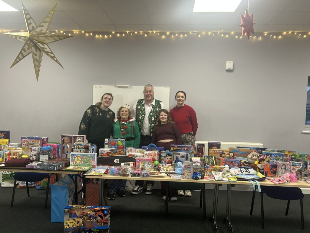 Members of Lyde Community Hub standing behind a table piled high with children's toys and gifts, decorated with Christmas lights and stars, gathered to support local families at Christmas.
