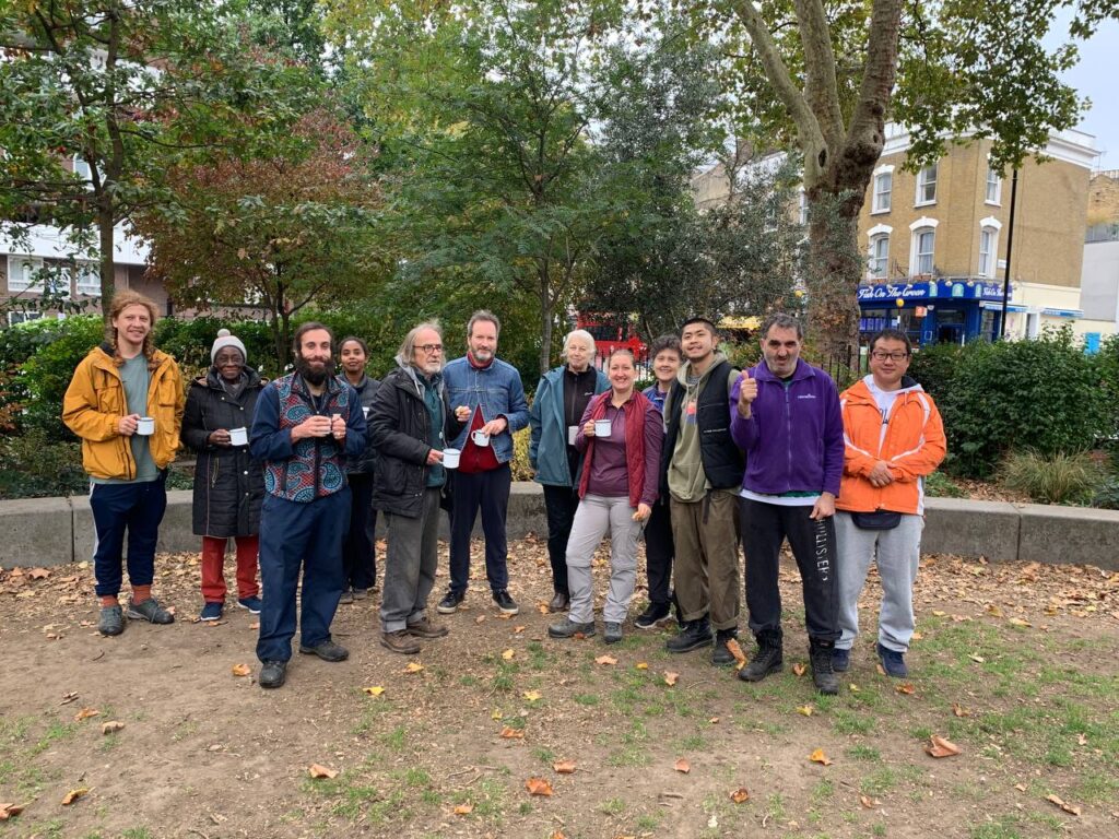 A group of around twelve volunteers and participants with additional needs stand together as part of The Garden Classroom smiling in Newington Green, Islington, dressed in warm autumn clothing and holding mugs of tea.