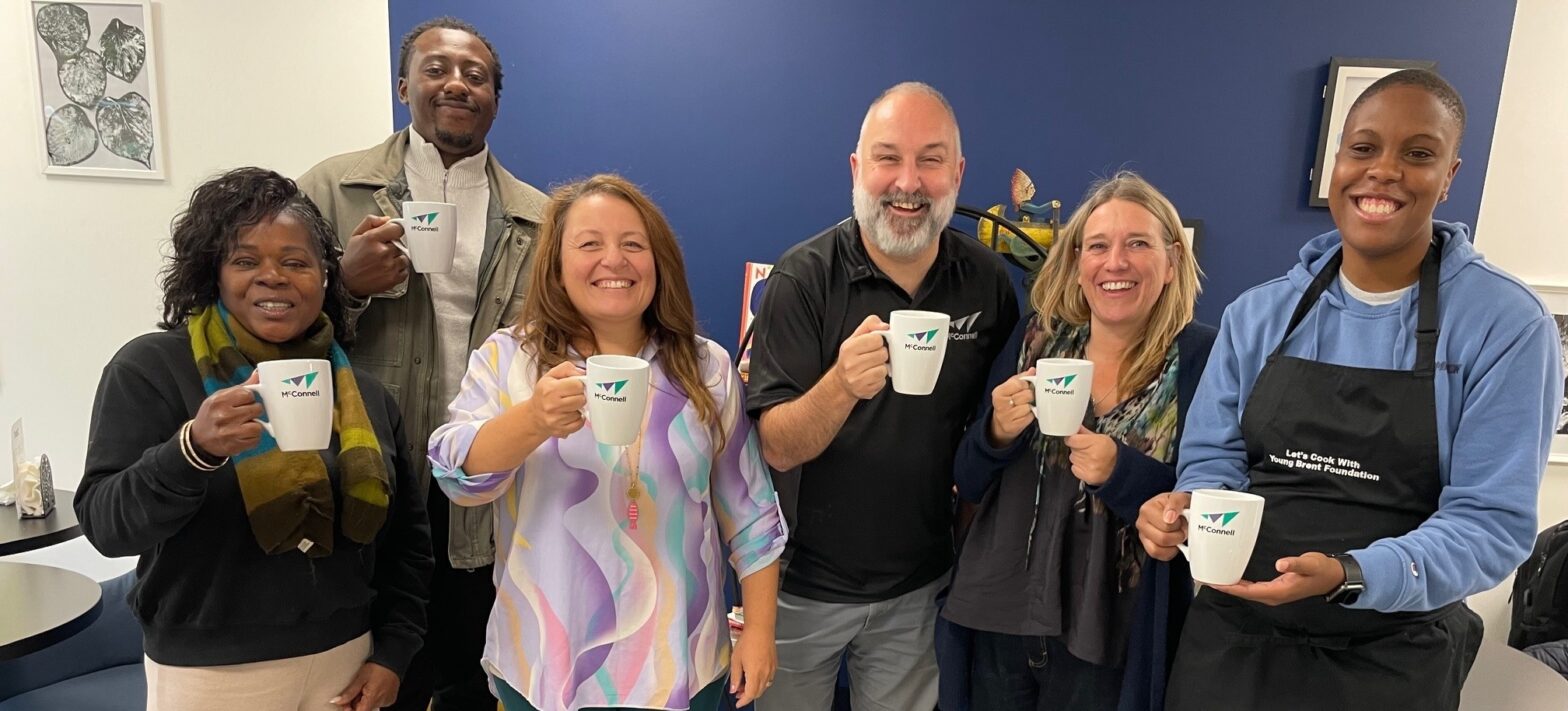 Julian Sanz, Head of Social Value at McConnell, and five team members from the Young Brent Foundation smile while holding donated coffee mugs for their new project - a coffee shop.