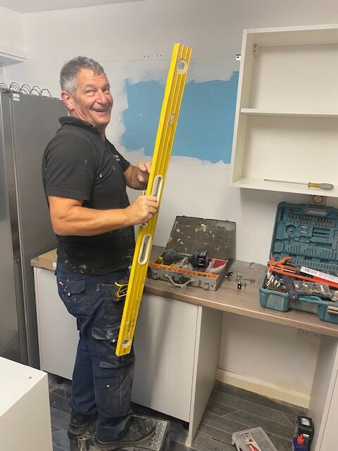 A man in a black polo shirt and dark work trousers stands on a small step stool in a kitchen under renovation. He is smiling and holding a long, yellow spirit level vertically. In the background, a section of the wall is painted light blue, and various tools—including open toolboxes, a drill, and a sealant gun—are scattered across a wooden countertop and open white shelving units.