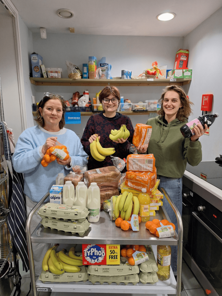 Three volunteers for the night shelter, Hackney Doorways, are photographed showcasing the essential food items including milk, eggs and bananas from three weekly grocery shops provided by supplier of the month, Alphatrack.