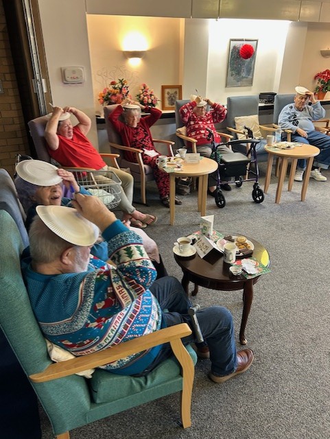 A group of older adults in Christmas jumpers laughing and balancing paper plates on their heads during a festive celebration in a care or community lounge setting in Basingstoke.