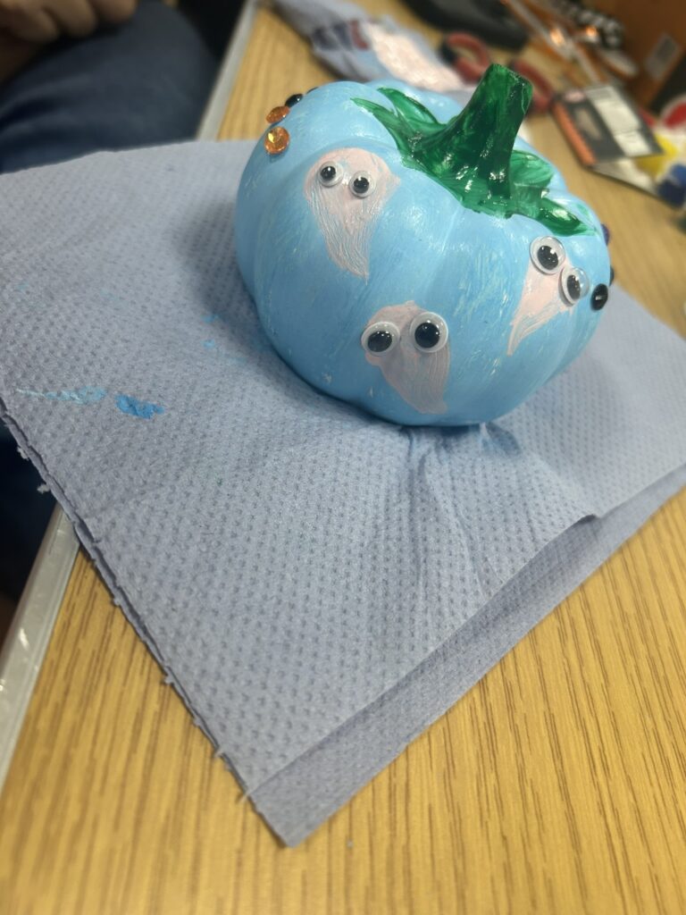 A small pumpkin painted light blue and decorated with googly eyes and orange gem stickers, resting on a blue paper towel on a wooden table, made during a Halloween craft session for children at Lyde Green Community Hub.